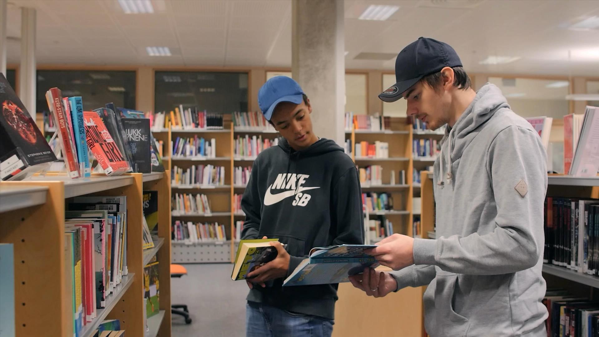 Foto av to elever i biblioteket. Reoler med bøker i bakgrunn
